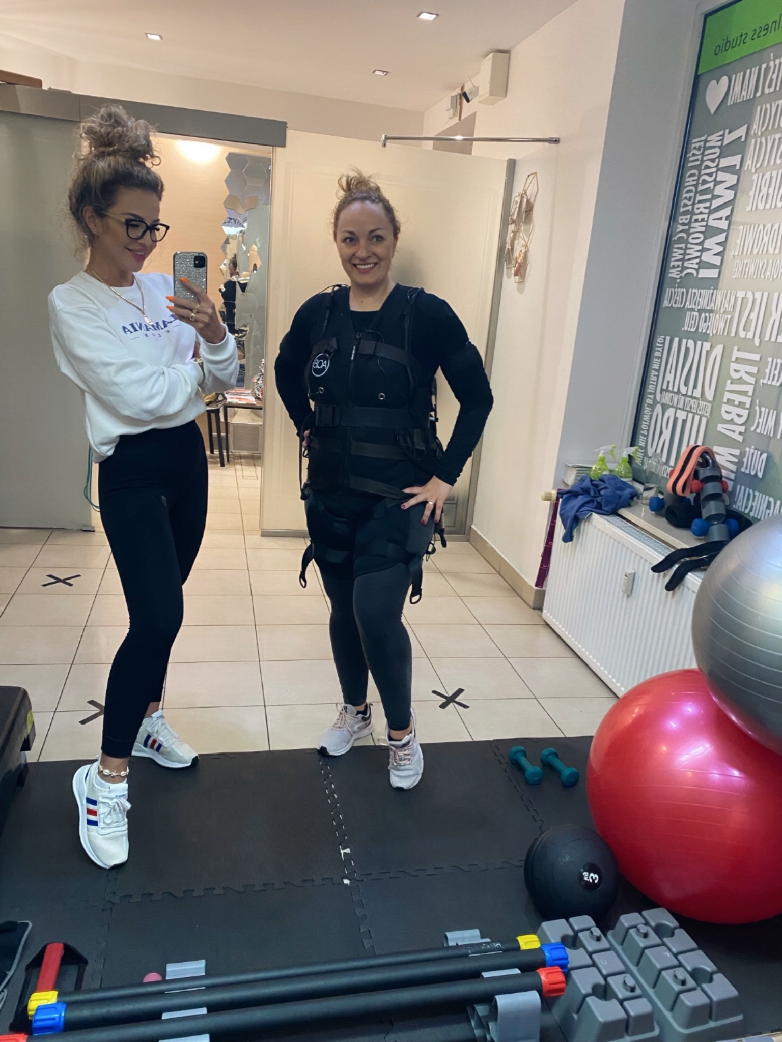 Kobieta w czarnym stroju do elektrostymulacji EMS pozuje z trenerką robiącą selfie w siłowni, na podłodze mata, w tle piłki do ćwiczeń, hantle i ściana z napisem.