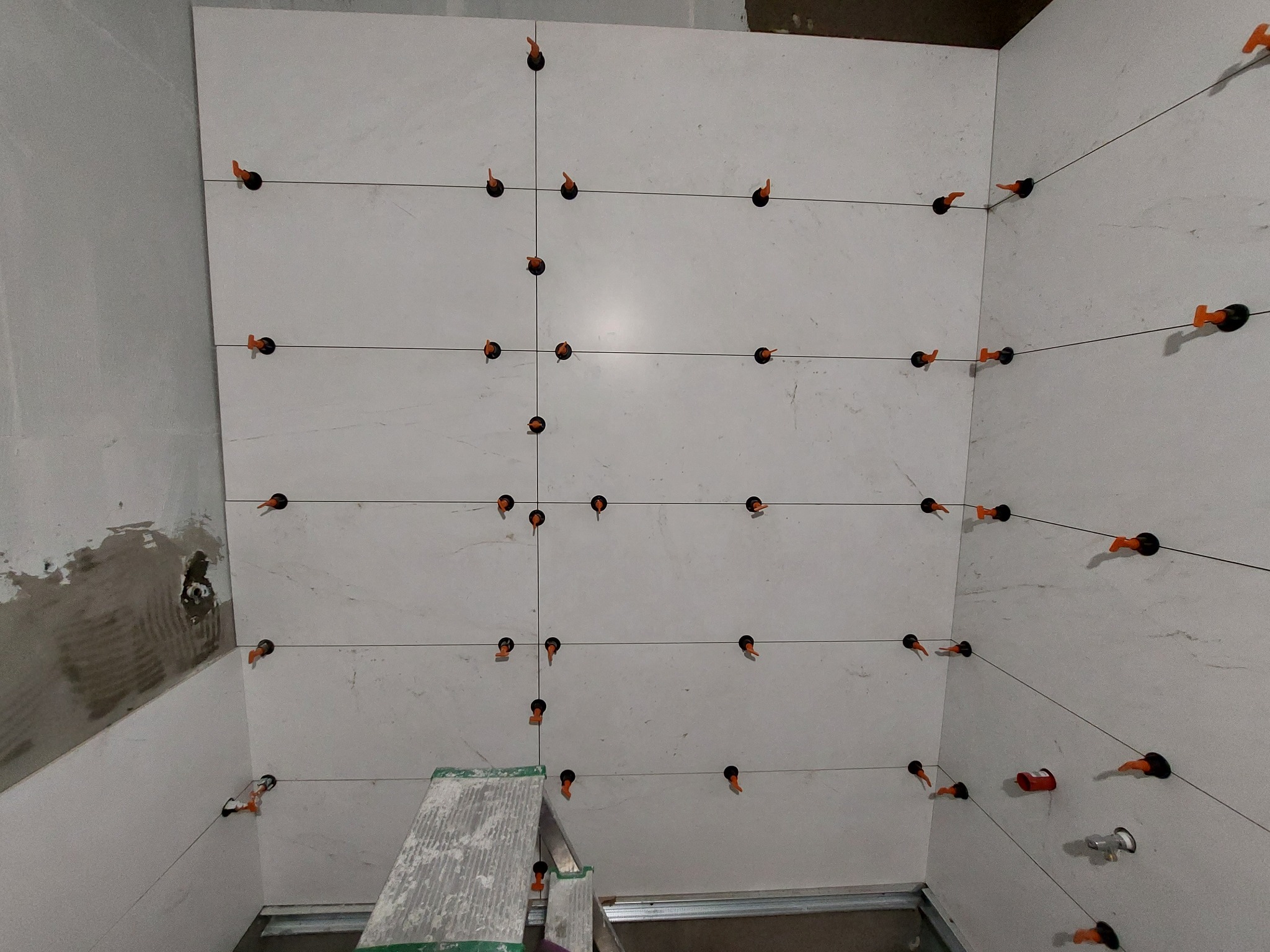 Ściana w trakcie układania białych płytek z użyciem systemu poziomowania, widoczne czarne klipsy i pomarańczowe kliny, fragment drabiny w dolnej części kadru.