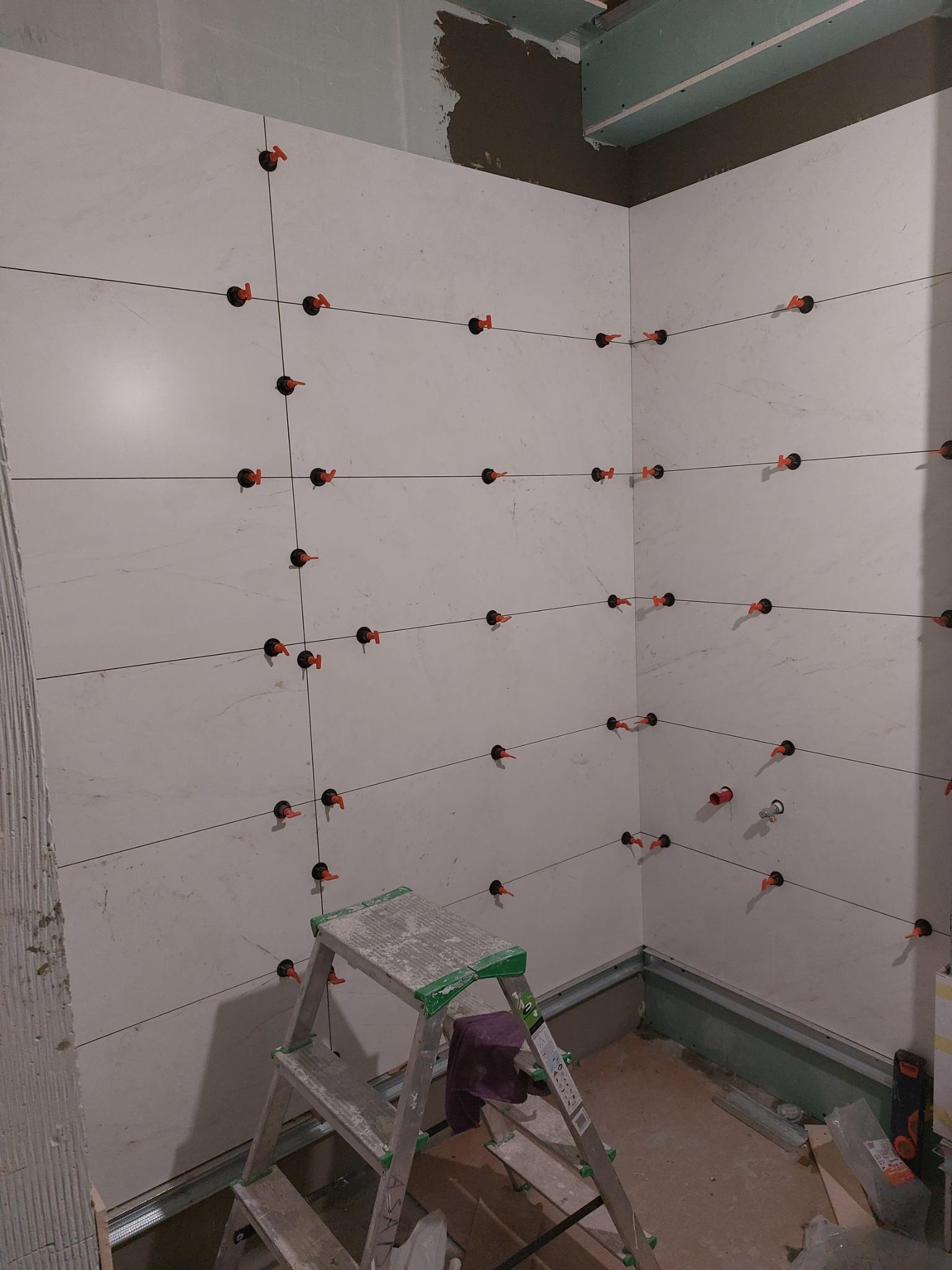 Układanie jasnych płytek w łazience z użyciem systemu poziomowania, widoczna drabina aluminiowa i elementy konstrukcyjne pod zabudowę