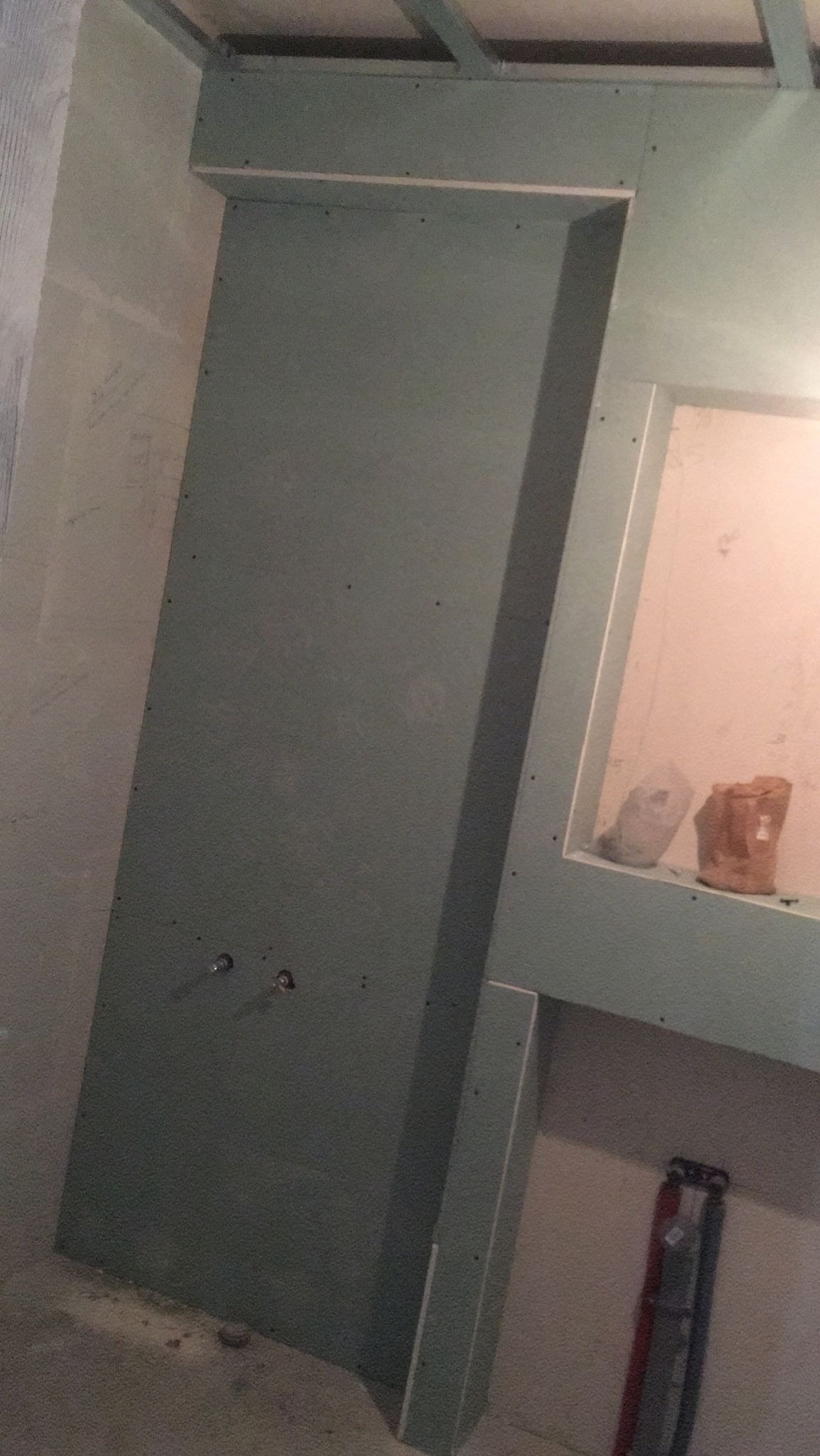 Konstrukcja z płyt gipsowo-kartonowych w trakcie montażu, widoczne rury instalacji wodnej i półka z materiałami budowlanymi.