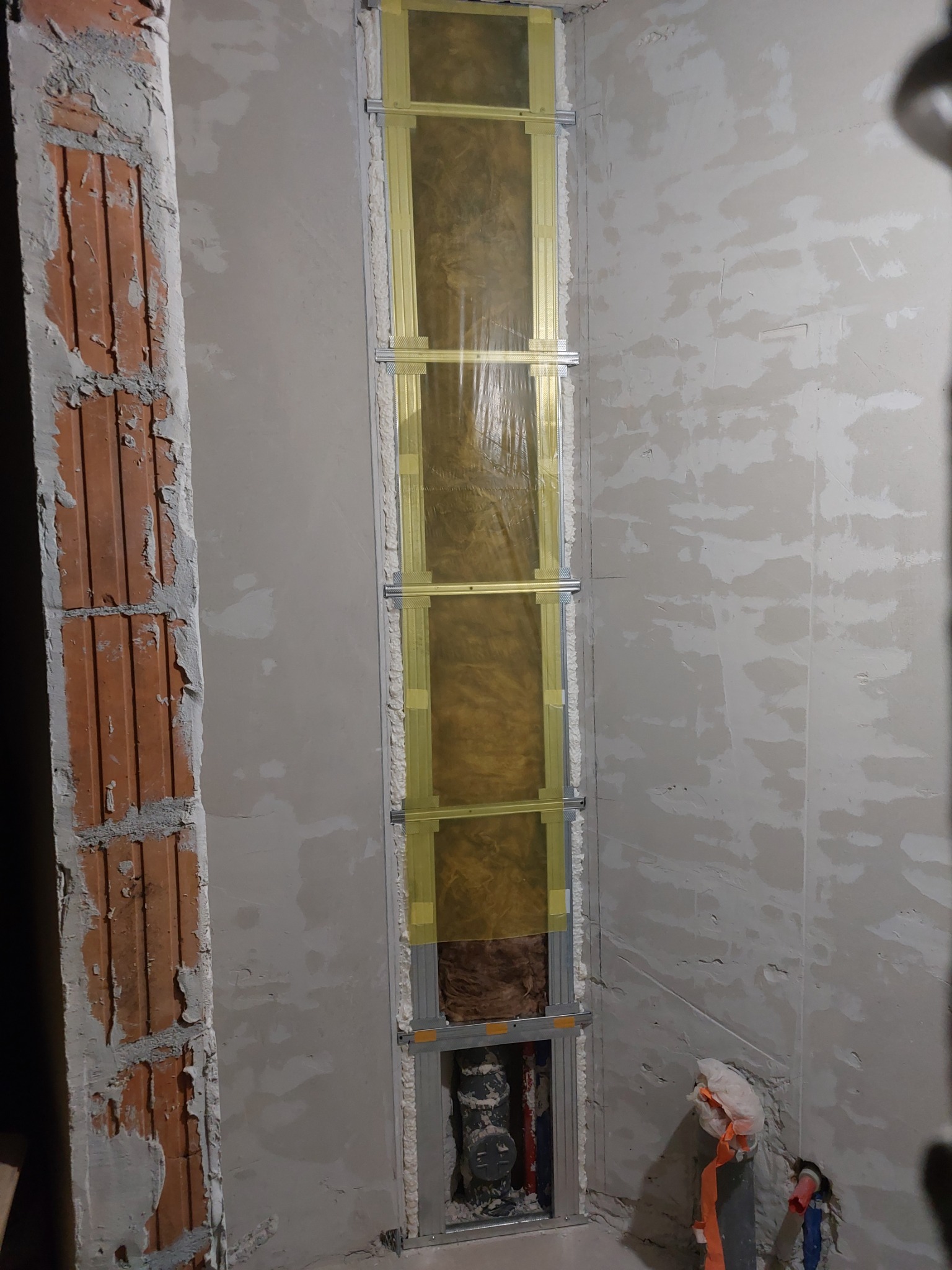 Wnęka w ścianie z widoczną instalacją hydrauliczną, ocieplona wełną mineralną i zabezpieczona folią, zamocowana na metalowym stelażu, obok fragment ściany z surową cegłą