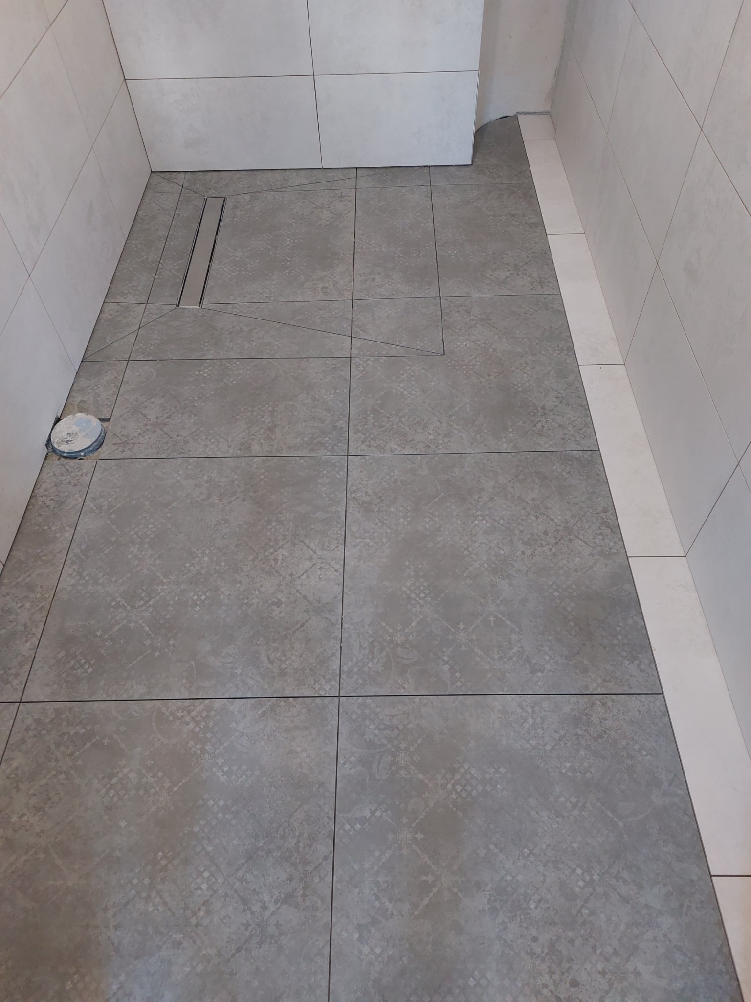 Ułożona szara płytka podłogowa w łazience z odpływem liniowym, widoczny fragment podłogi przed wejściem do kabiny prysznicowej, białe płytki ścienne.