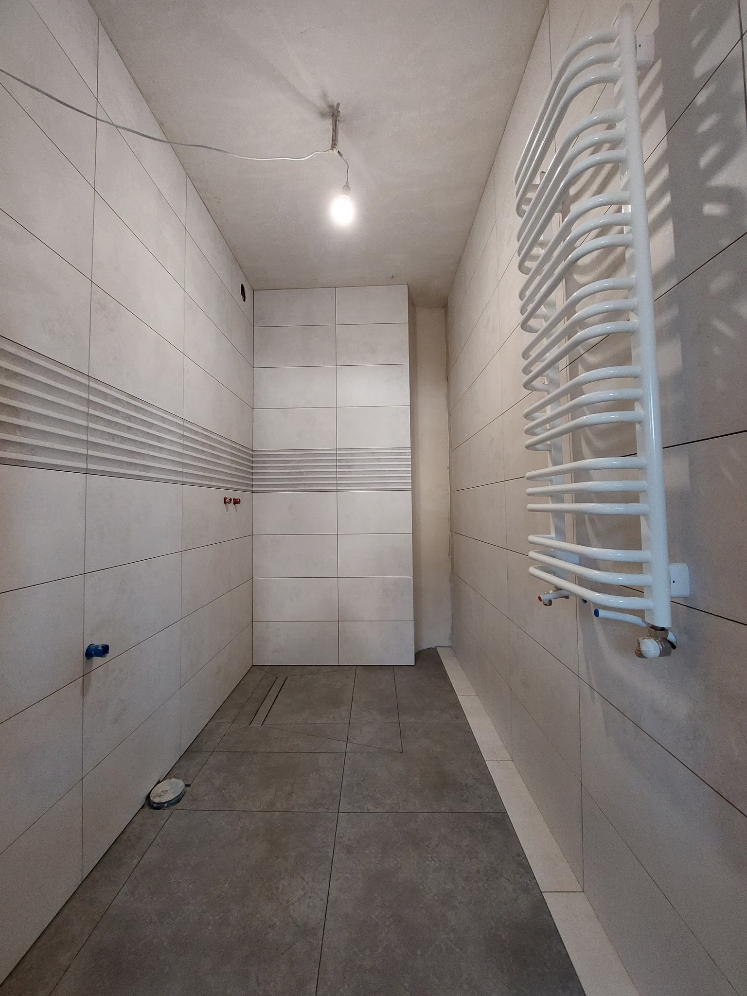 Wnętrze łazienki w trakcie prac wykończeniowych: ułożone jasne płytki ścienne z dekoracyjnym pasem, ciemne płytki podłogowe z odpływem liniowym, biały grzejnik drabinkowy, surowa żarówka zwisająca...