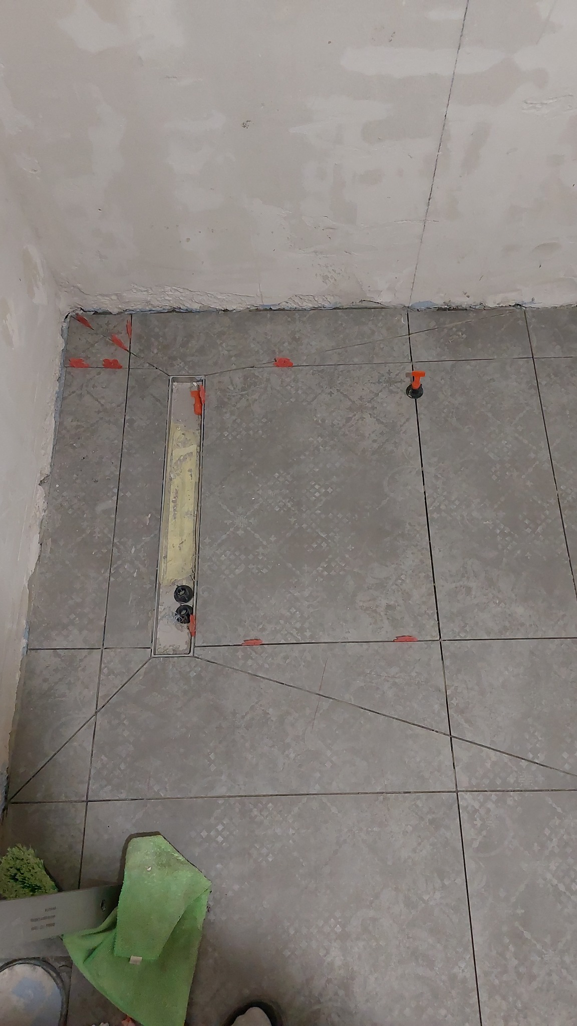 Ułożone szare płytki podłogowe z odpływem liniowym, widoczne pomarańczowe krzyżyki dystansowe, ściany przygotowane do dalszych prac, na podłodze leży zielona ściereczka.