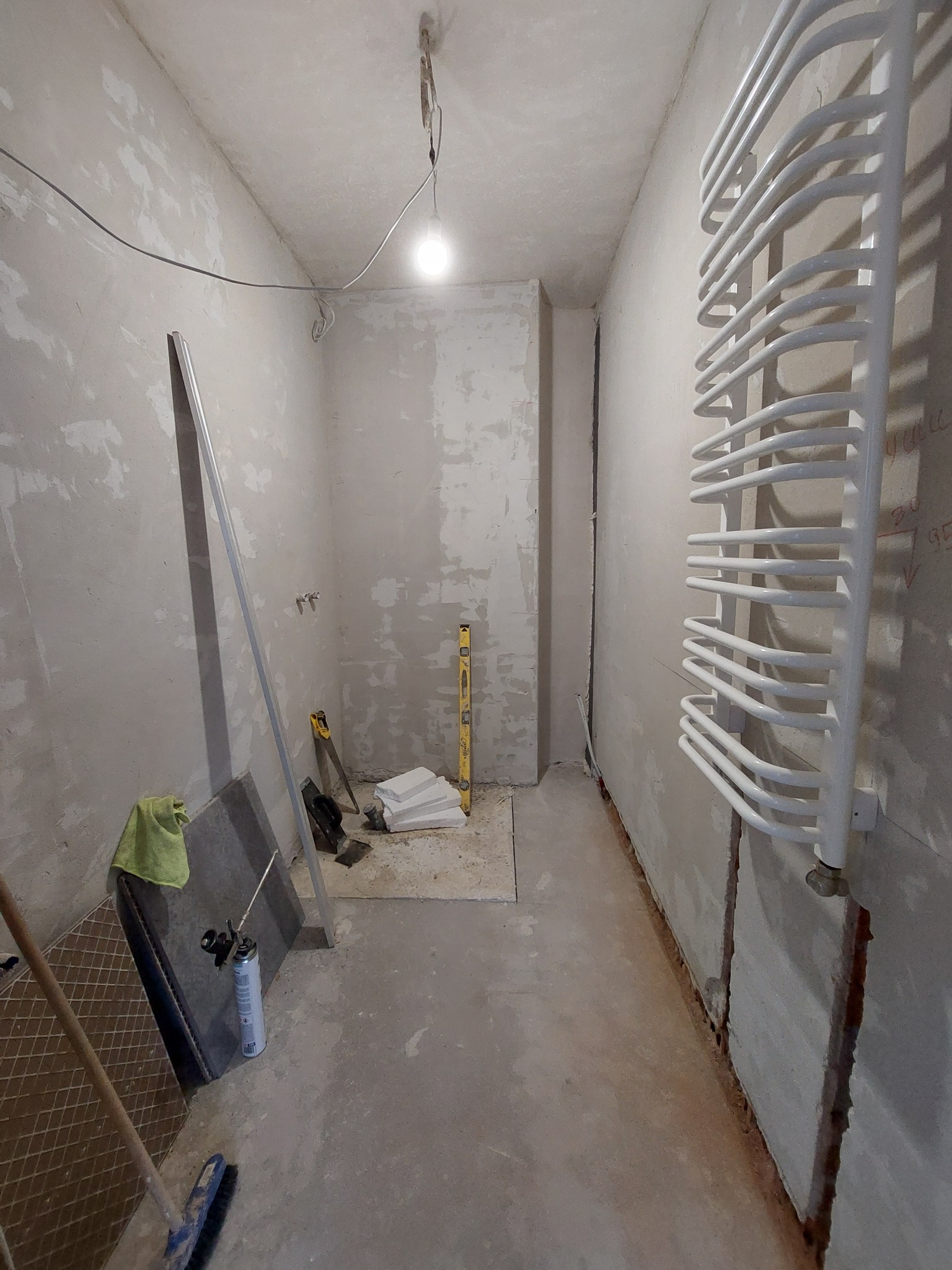 Wnętrze łazienki w trakcie remontu: surowe ściany z widocznymi śladami szpachlowania, biały grzejnik drabinkowy, narzędzia glazurnicze, poziomica i płytki podłogowe przygotowane do ułożenia.