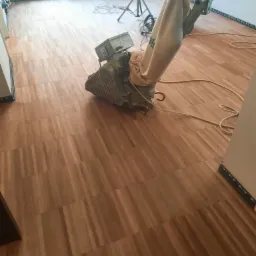 Szlifowanie drewnianej podłogi parkietowej maszyną z workiem na pył, widoczne kable i oświetlenie pomocnicze, ściany pomalowane na biało.