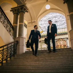 Dwóch mężczyzn w garniturach schodzi po schodach w budynku o architekturze klasycznej we Wrocławiu.