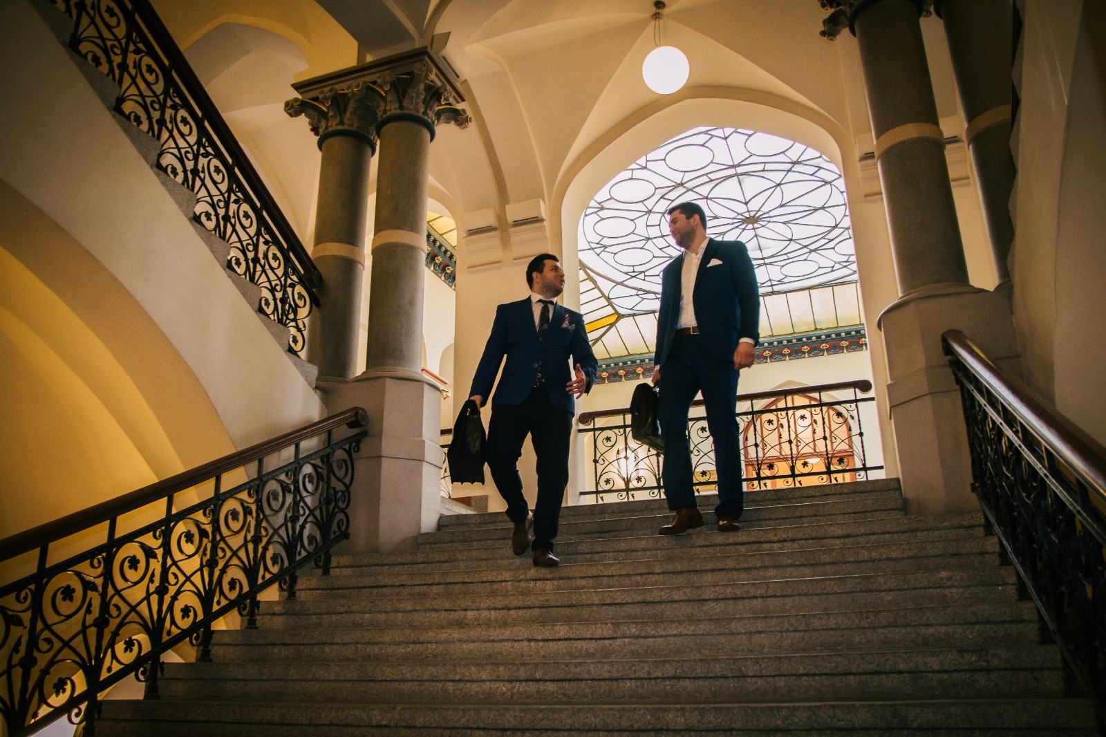 Dwóch mężczyzn w garniturach schodzi po schodach w budynku o architekturze klasycznej we Wrocławiu.