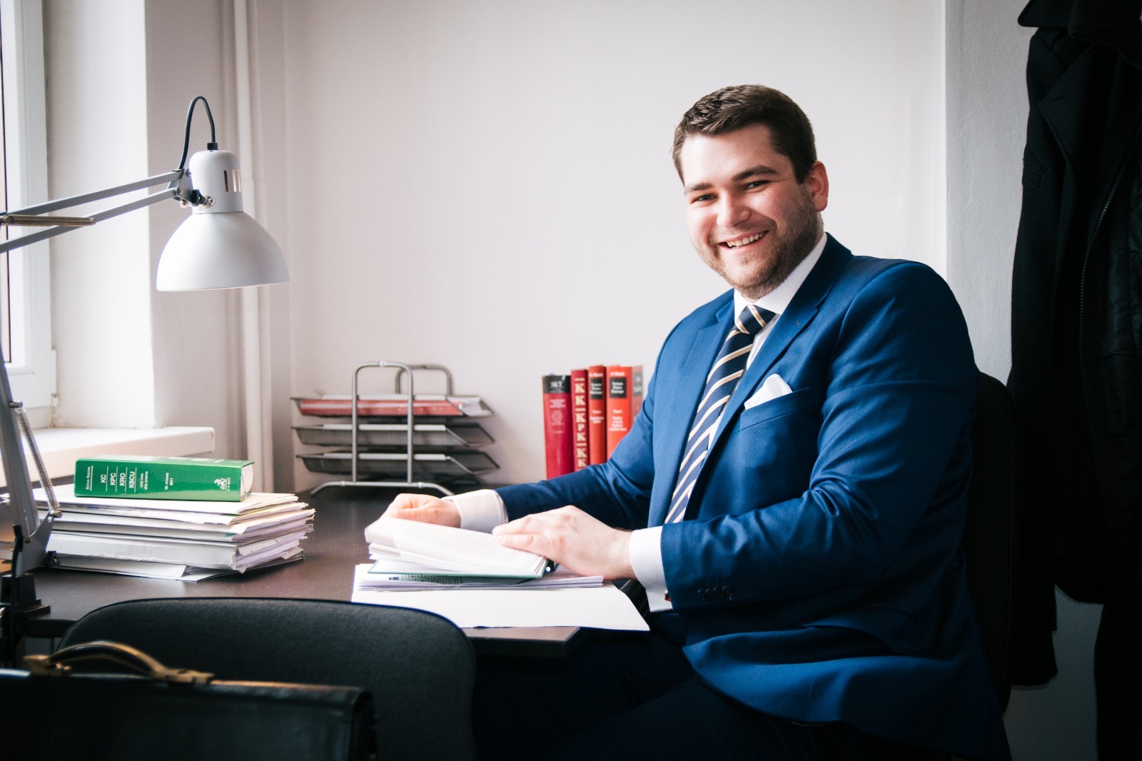 Uśmiechnięty mężczyzna w niebieskim garniturze siedzi za biurkiem z dokumentami, książkami prawniczymi i lampką, spoglądając w kamerę.