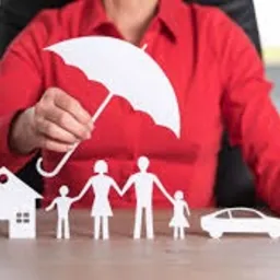 Osoba w czerwonej koszuli trzyma biały, papierowy parasol nad wyciętymi z papieru sylwetkami rodziny, domu i samochodu, leżącymi na drewnianym stole.