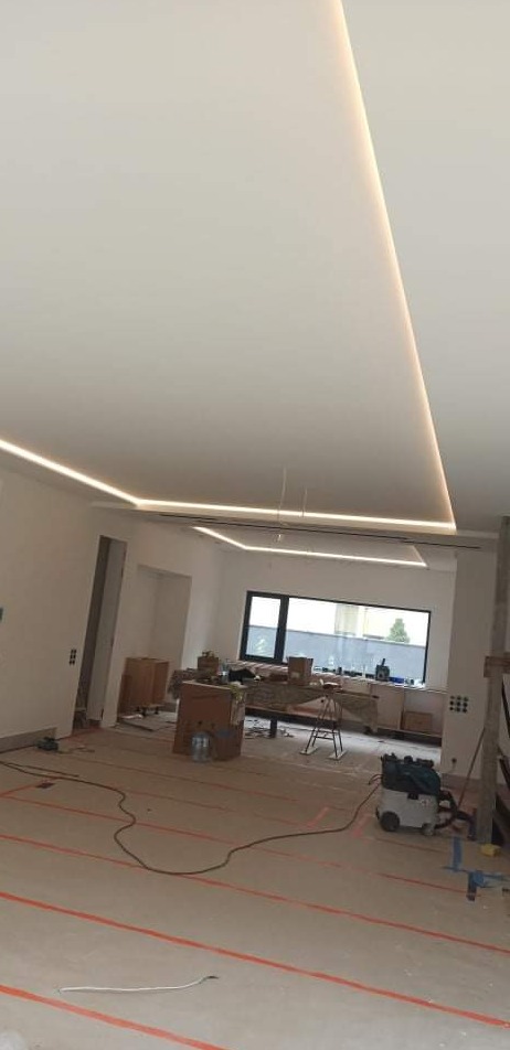 Wnętrze w trakcie prac wykończeniowych: widoczny sufit z oświetleniem LED, podłoga z zaznaczonymi liniami, kartony i narzędzia w tle.
