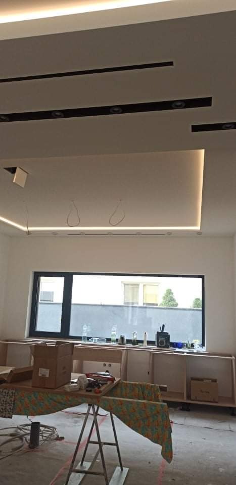 Sufit podwieszany z oświetleniem LED w trakcie montażu, widoczne kable, okno z widokiem na zewnątrz, w tle meble w stanie surowym.