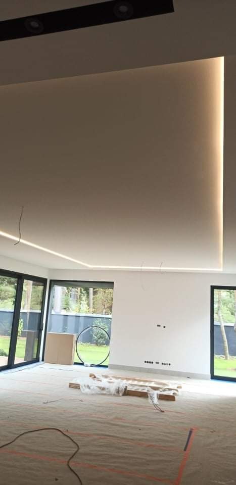 Wnętrze w trakcie wykańczania, z widocznym podwieszanym sufitem z oświetleniem LED, zabezpieczoną podłogą i dużymi oknami z widokiem na zieleń.