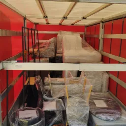 Wnętrze czerwonej furgonetki wypełnionej meblami owiniętymi folią stretch, zabezpieczonymi na czas transportu, widoczne krzesła, stół i elementy wyposażenia salonu.