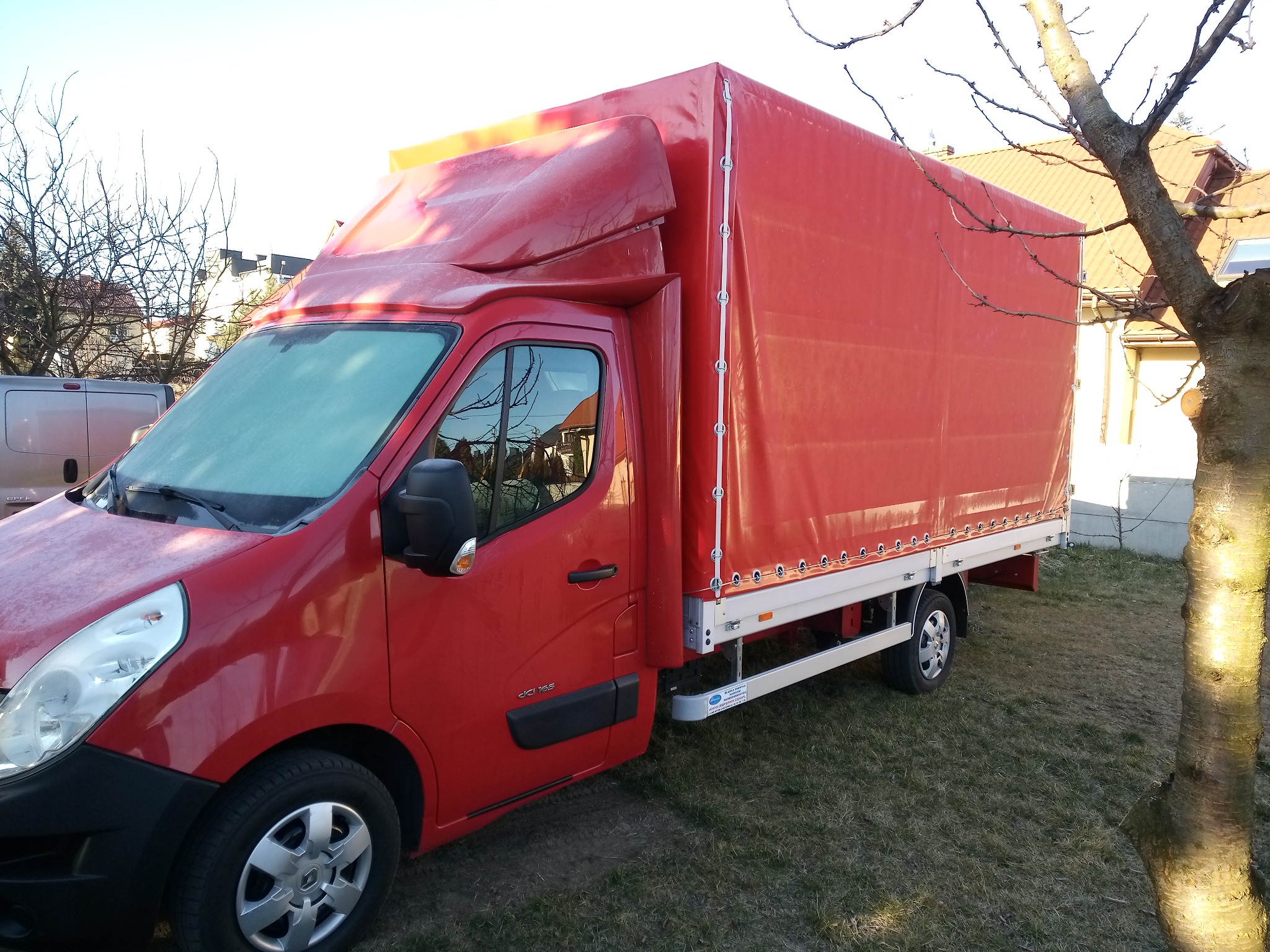 Czerwony samochód dostawczy z plandeką na trawniku przed domem, widoczne krople wody na karoserii.