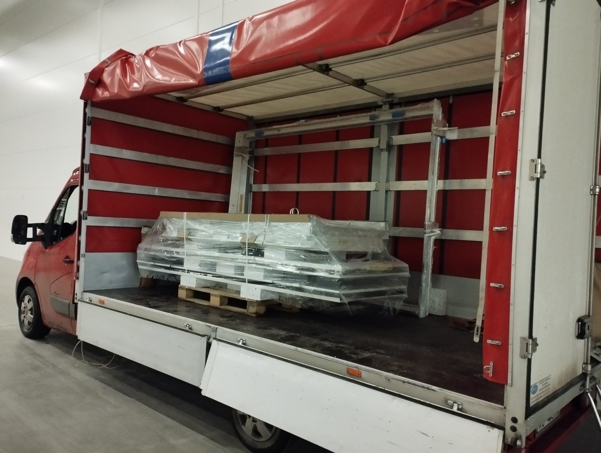 Czerwony bus dostawczy z otwartą przestrzenią ładunkową, w której znajdują się zabezpieczone folią okna na drewnianej palecie.