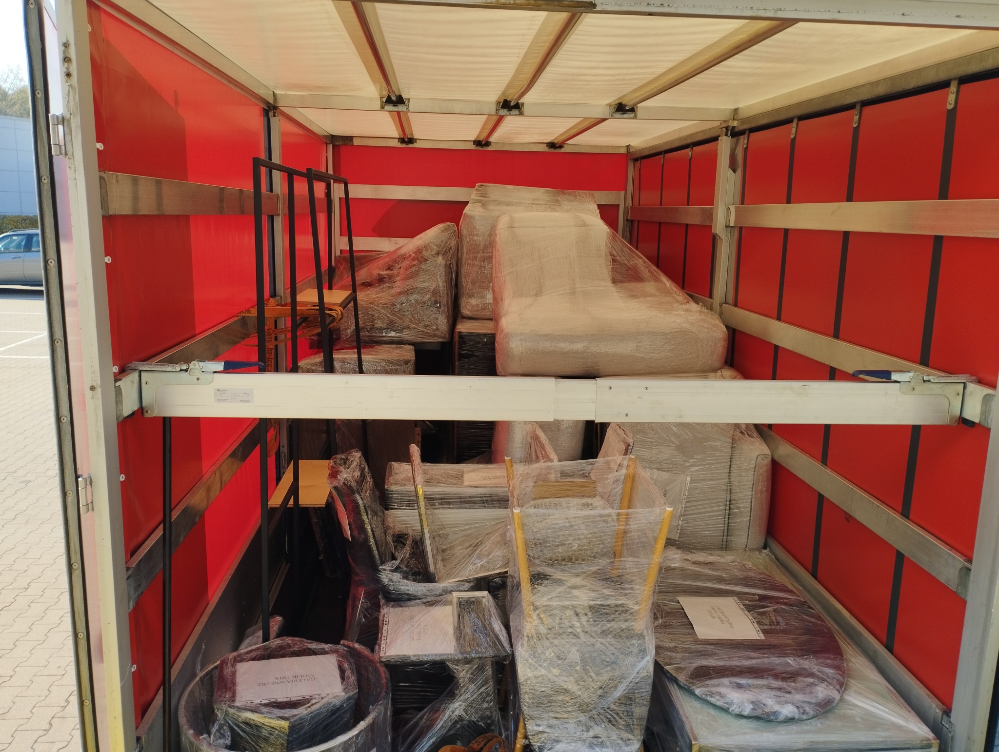 Wnętrze czerwonej furgonetki wypełnionej meblami owiniętymi folią stretch, zabezpieczonymi na czas transportu, widoczne krzesła, stół i elementy wyposażenia salonu.