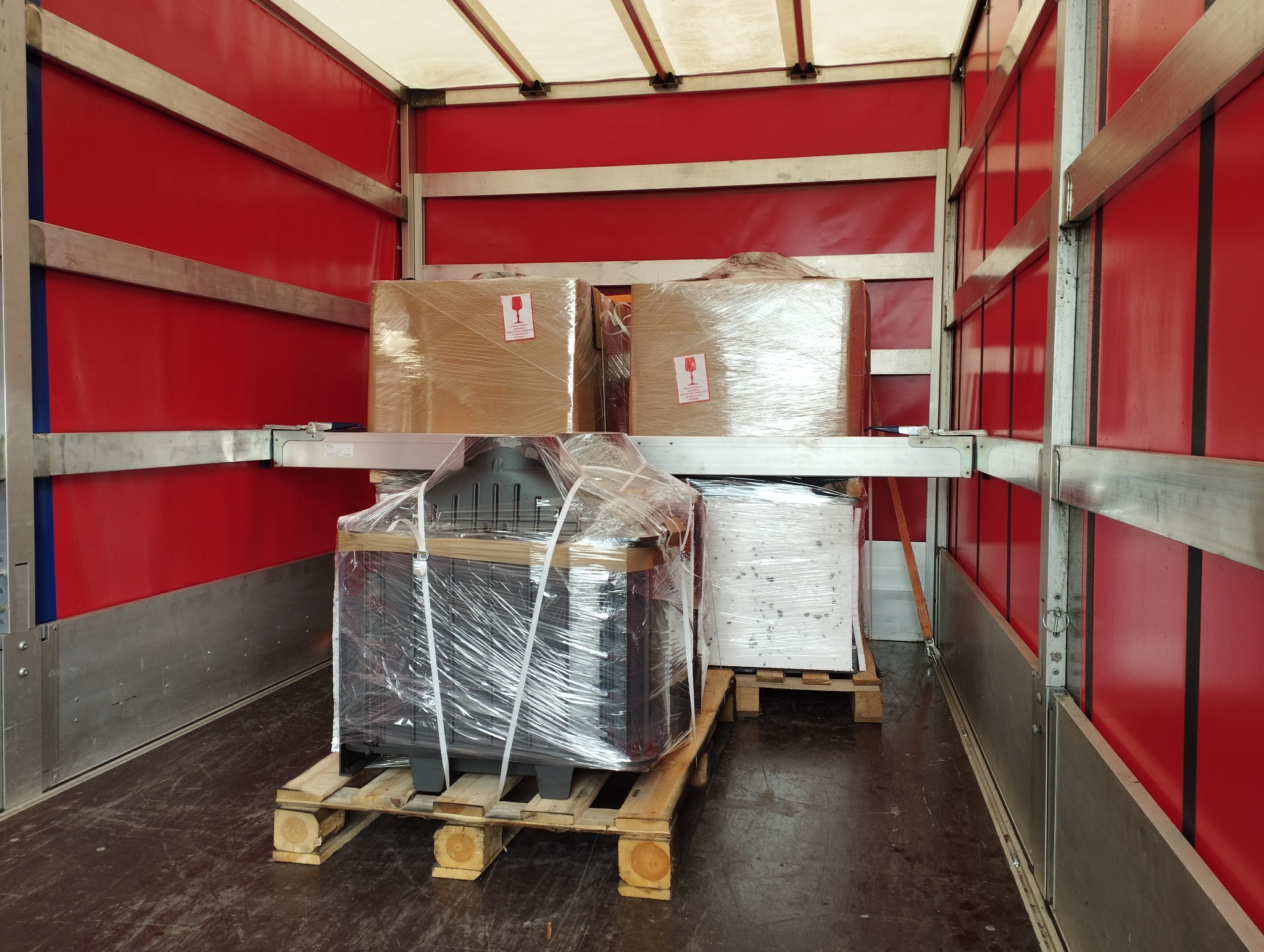 Wnętrze furgonetki z czerwonymi ścianami, załadowanej paletami z zapakowanymi w folię przedmiotami, w tym kartonami z naklejkami 'Fragile' i czarnym piecem kominkowym.