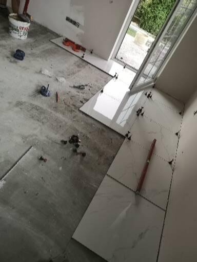 Układanie białych płytek podłogowych w pomieszczeniu z oknem, widoczne narzędzia i poziomica, klej w wiadrze w rogu pomieszczenia.