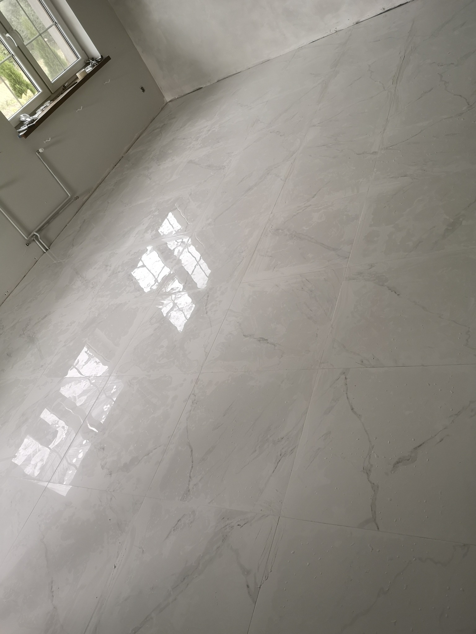 Jasne, błyszczące płytki podłogowe imitujące marmur w nowym wnętrzu w Słubicach, z widocznym oknem i grzejnikiem na ścianie.