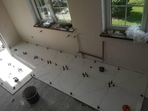 Ułożone białe płytki podłogowe z czarnymi krzyżykami dystansowymi w jasnym pomieszczeniu z dwoma oknami, widoczny kubeł i narzędzia.