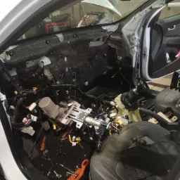 Wnętrze białego samochodu w trakcie demontażu deski rozdzielczej i elementów elektrycznych, widoczne liczne kable, zdemontowana tapicerka drzwi i elementy układu kierowniczego.