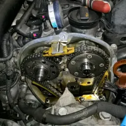 Otwarty silnik samochodowy z widocznymi łańcuchami rozrządu, z użyciem narzędzia pomiarowego, sugerujący precyzyjną regulację lub naprawę. Widoczne ślady oleju. Dolna część silnika zakryta szmatką.