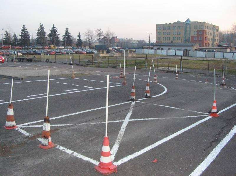 Plac manewrowy z wyznaczonymi liniami i ustawionymi pachołkami z białymi tyczkami, w tle budynek i zaparkowane samochody.