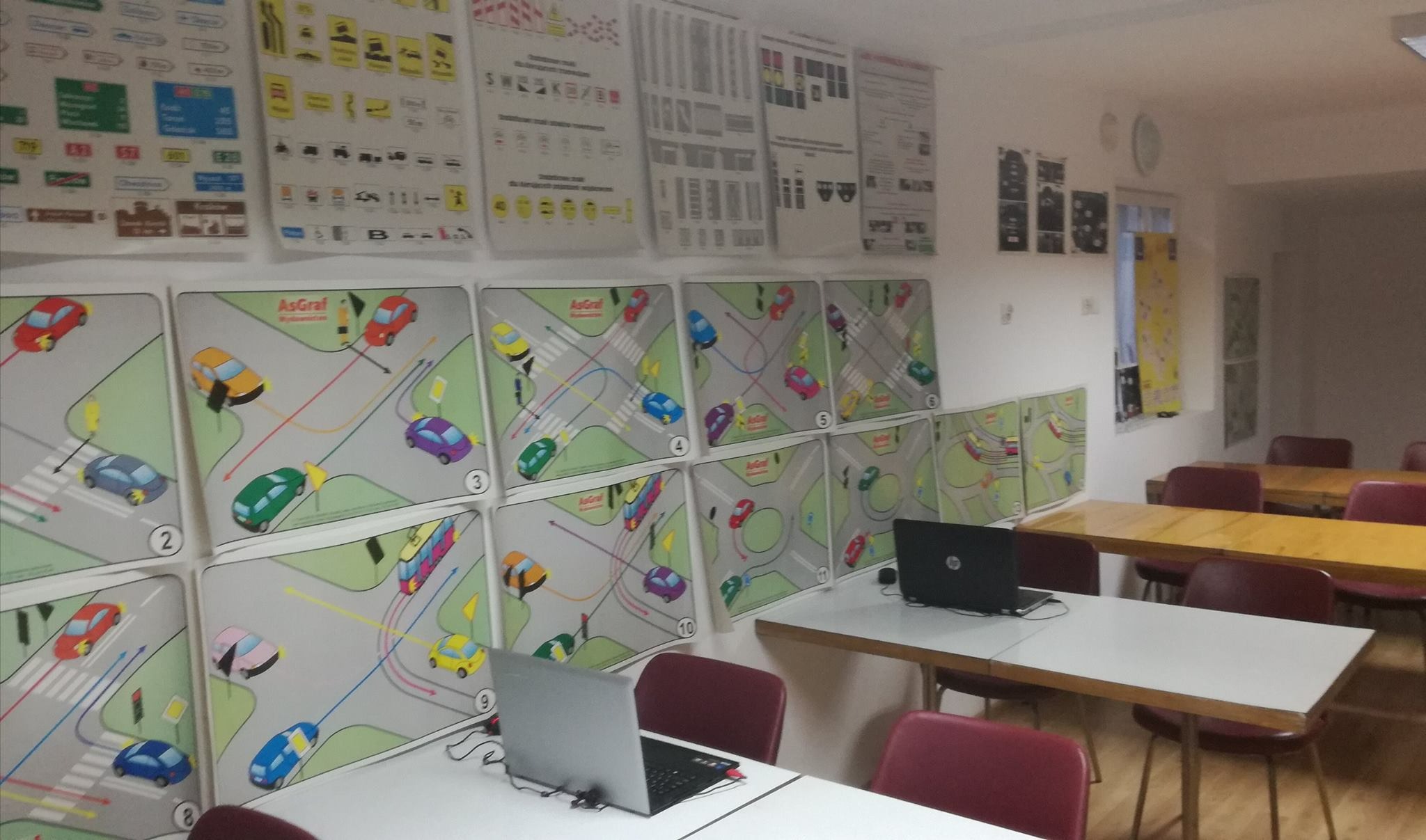 Sala dydaktyczna z planszami schematów skrzyżowań i znaków drogowych na ścianach, biurkami i laptopami, przygotowana do zajęć teoretycznych.
