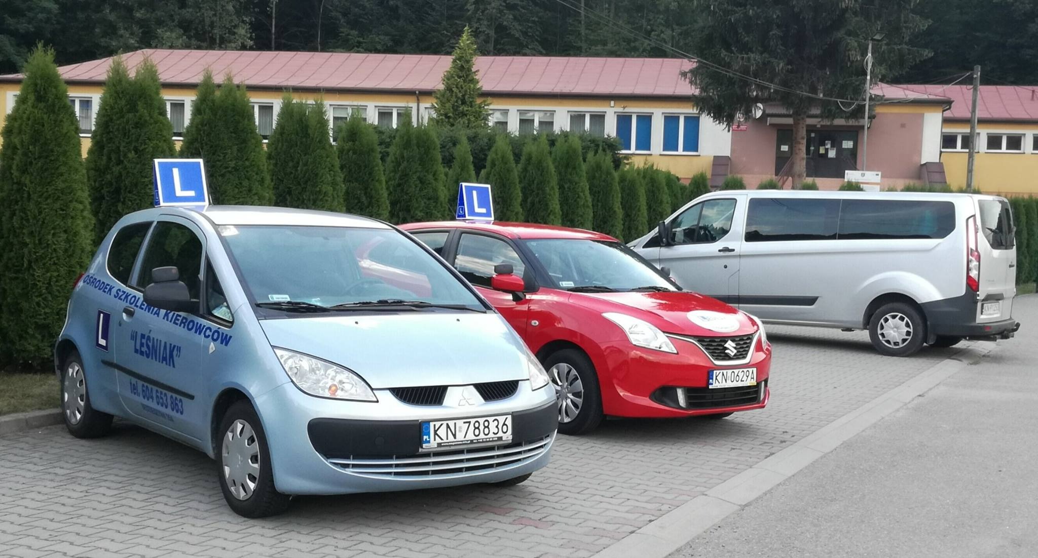 Trzy pojazdy oznakowane literą 'L' zaparkowane przed budynkiem: błękitny samochód z napisem 'Ośrodek Szkolenia Kierowców Leśniak', czerwony samochód i srebrny bus.