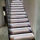 Podświetlone schody z czarnymi poręczami i białym światłem LED, widok z dołu, częściowo zabezpieczone taśmą malarską, w tle widoczne drzwi.