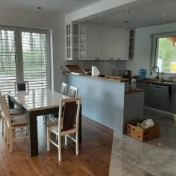 Jasna kuchnia z wyspą i jadalnią, parkiet w jodełkę, ceglana ściana, okno z żaluzjami.