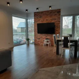 Jasne wnętrze z drewnianą podłogą, ceglaną ścianą z telewizorem i stołem jadalnym z krzesłami. Widoczne rolety w oknach i oświetlenie sufitowe.