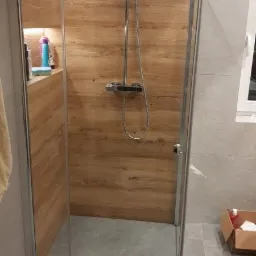 Kabina prysznicowa z drewnopodobną ścianą, szklane drzwi z chromowanymi zawiasami, deszczownica i półka z kosmetykami, szara podłoga, oświetlenie LED w suficie.