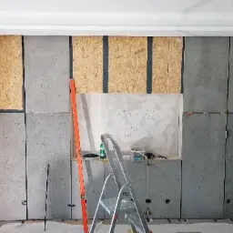 Ściana w trakcie obkładania płytami OSB i szarymi płytami, widoczna drabina i poziomica, przygotowania do dalszych prac wykończeniowych.