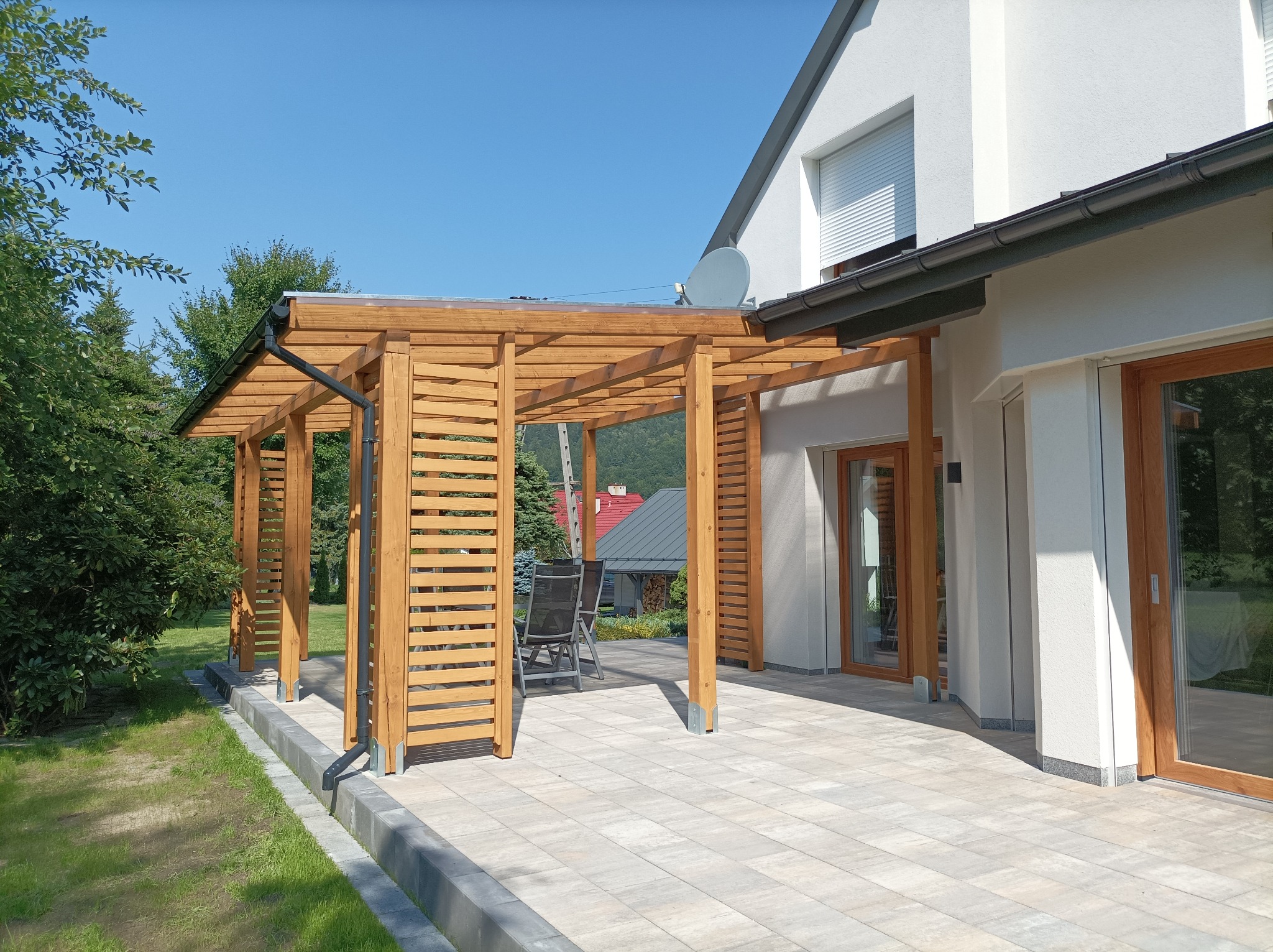 Drewniana altana z lamelami i przezroczystym dachem, z rynną odprowadzającą wodę, przylegająca do nowoczesnego domu z jasną elewacją i kostką brukową