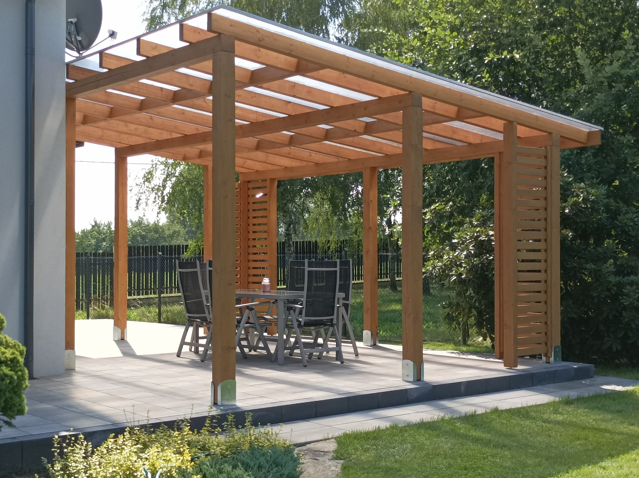 Zadaszona konstrukcja z drewna z transparentnym dachem, wyposażona w stół i krzesła na szarym, wyłożonym kostką tarasie, otoczona zielenią.