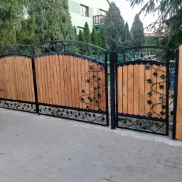 Drewniana brama wjazdowa z metalowymi elementami dekoracyjnymi w kształcie liści i winogron, widoczna częściowo biała karoseria samochodu, kostka brukowa i zieleń w tle.