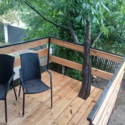 Drewniany taras z balustradą zintegrowany z drzewem, wyposażony w dwa czarne krzesła z plecionki, widok na ogród z zabawkami.