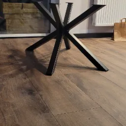 Surowa, czarna, metalowa podstawa stołu z ośmioma ramionami, widoczna na drewnianej podłodze w pomieszczeniu z grzejnikiem i oknem. W tle kartonowa torba.