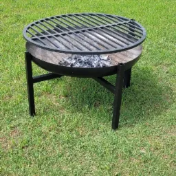 Metalowy grill ogrodowy z okrągłym rusztem i resztkami popiołu w palenisku, stojący na trawniku, w tle rząd tuj.