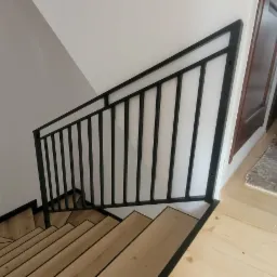 Wnętrze domu z drewnianymi schodami i czarną, metalową balustradą, widok z góry na dół, z fragmentem korytarza w tle.