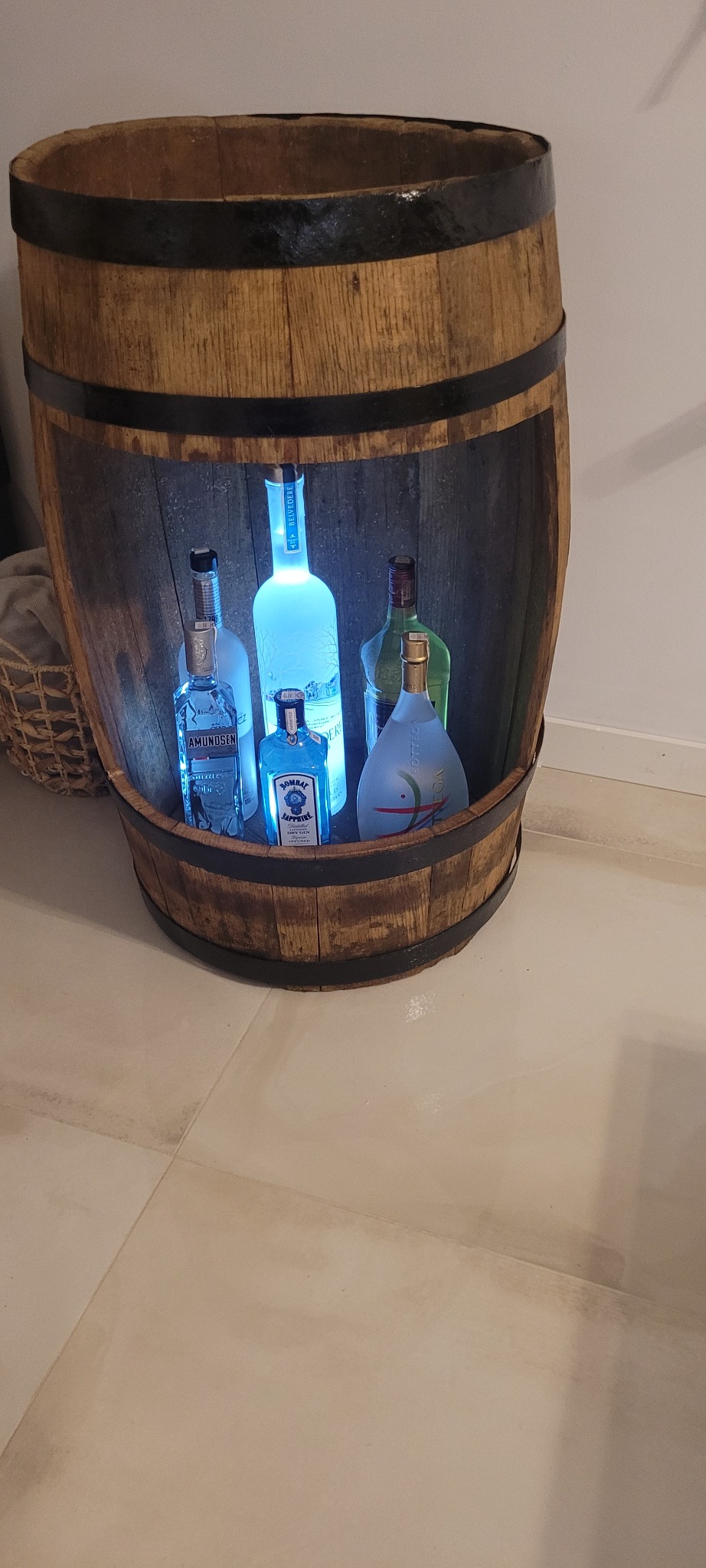 Drewniana beczka z wyciętym frontem, pełniąca funkcję barku, podświetlona od wewnątrz, eksponująca kolekcję markowych butelek alkoholu.