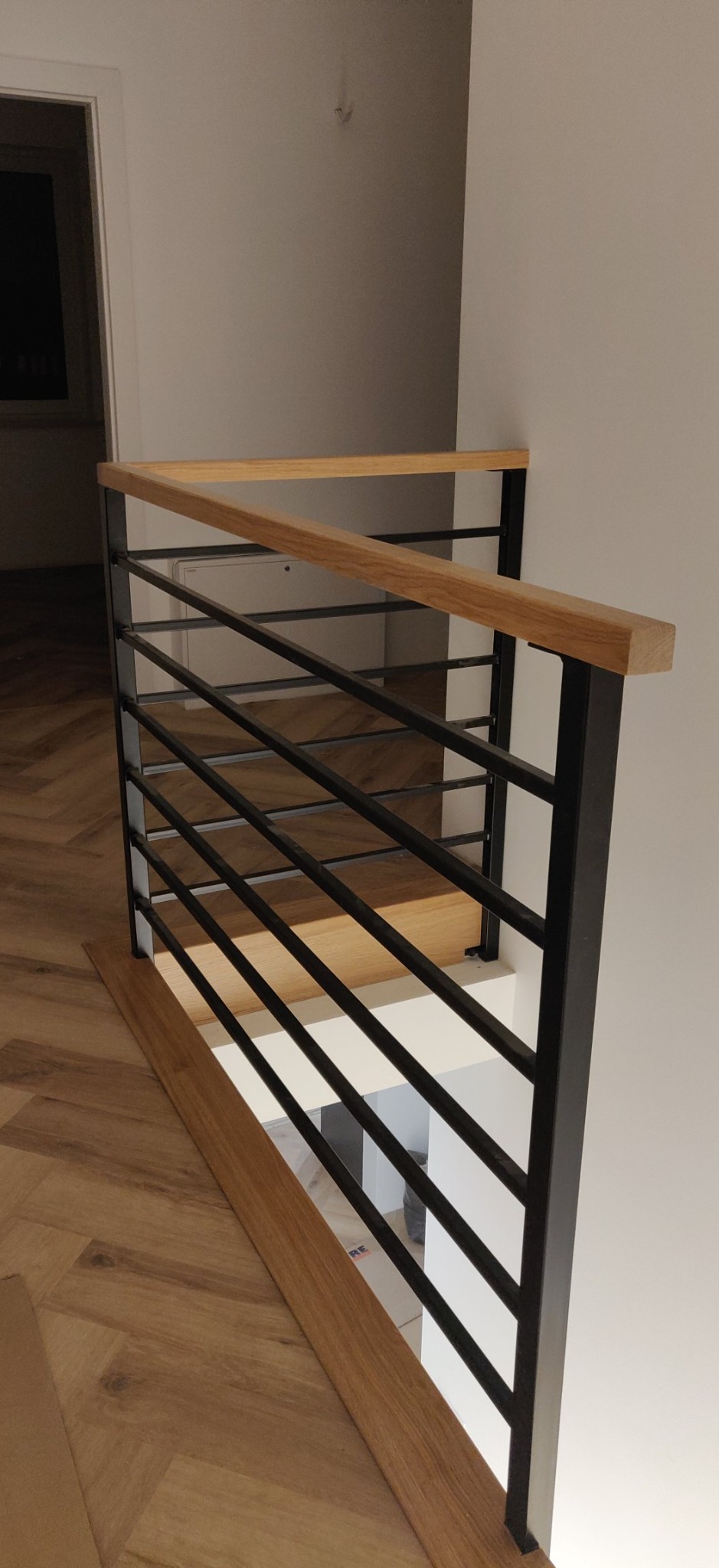 Nowoczesna balustrada schodowa z czarnych metalowych prętów i drewnianym poręczą, widok z góry na schody z parkietem w jodełkę.