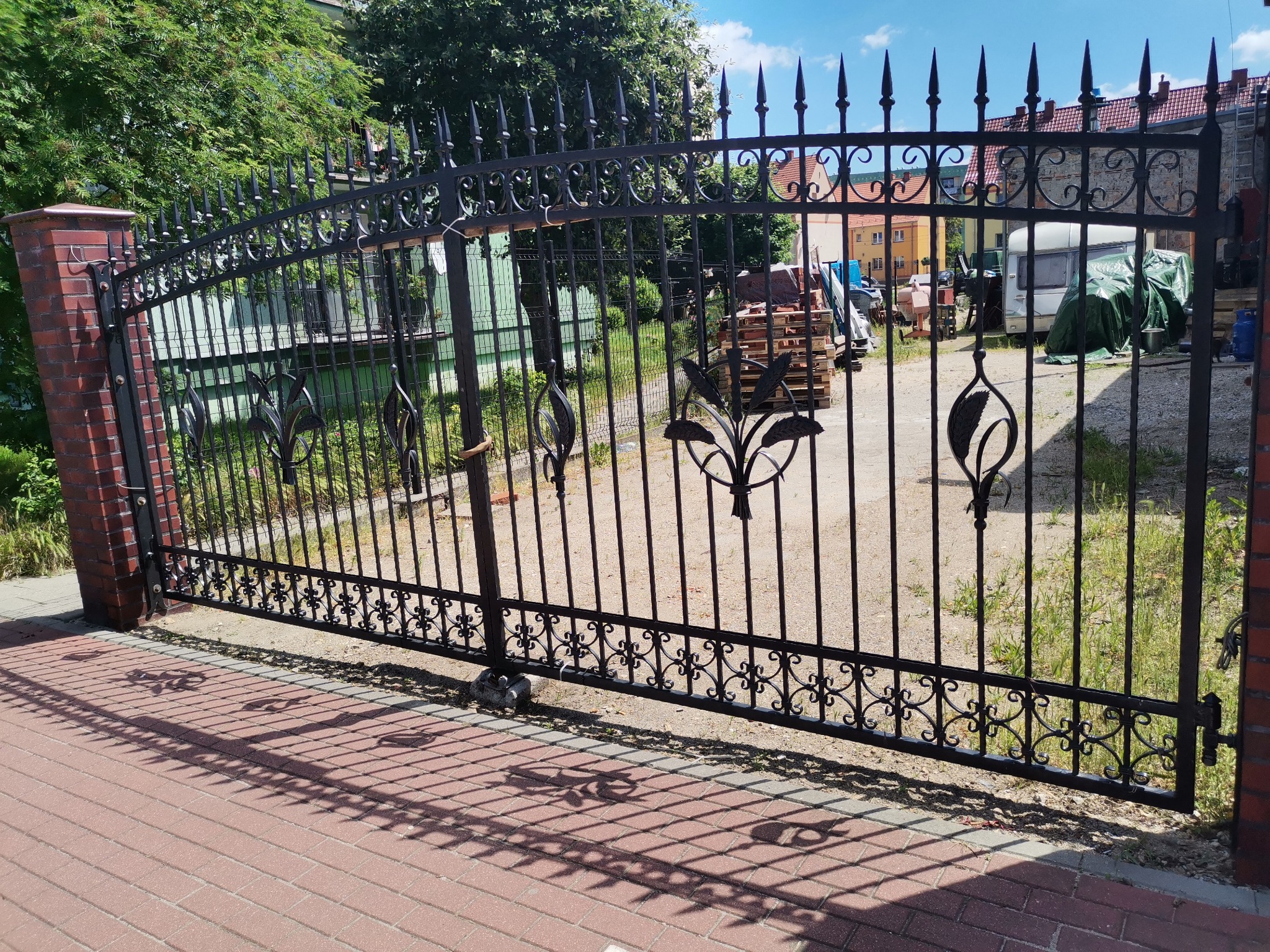 Otwarta, czarna brama wjazdowa z metalowymi prętami zakończonymi grotami, zdobiona motywem roślinnym, osadzona na ceglanym słupku, rzucająca cień na czerwoną kostkę brukową.