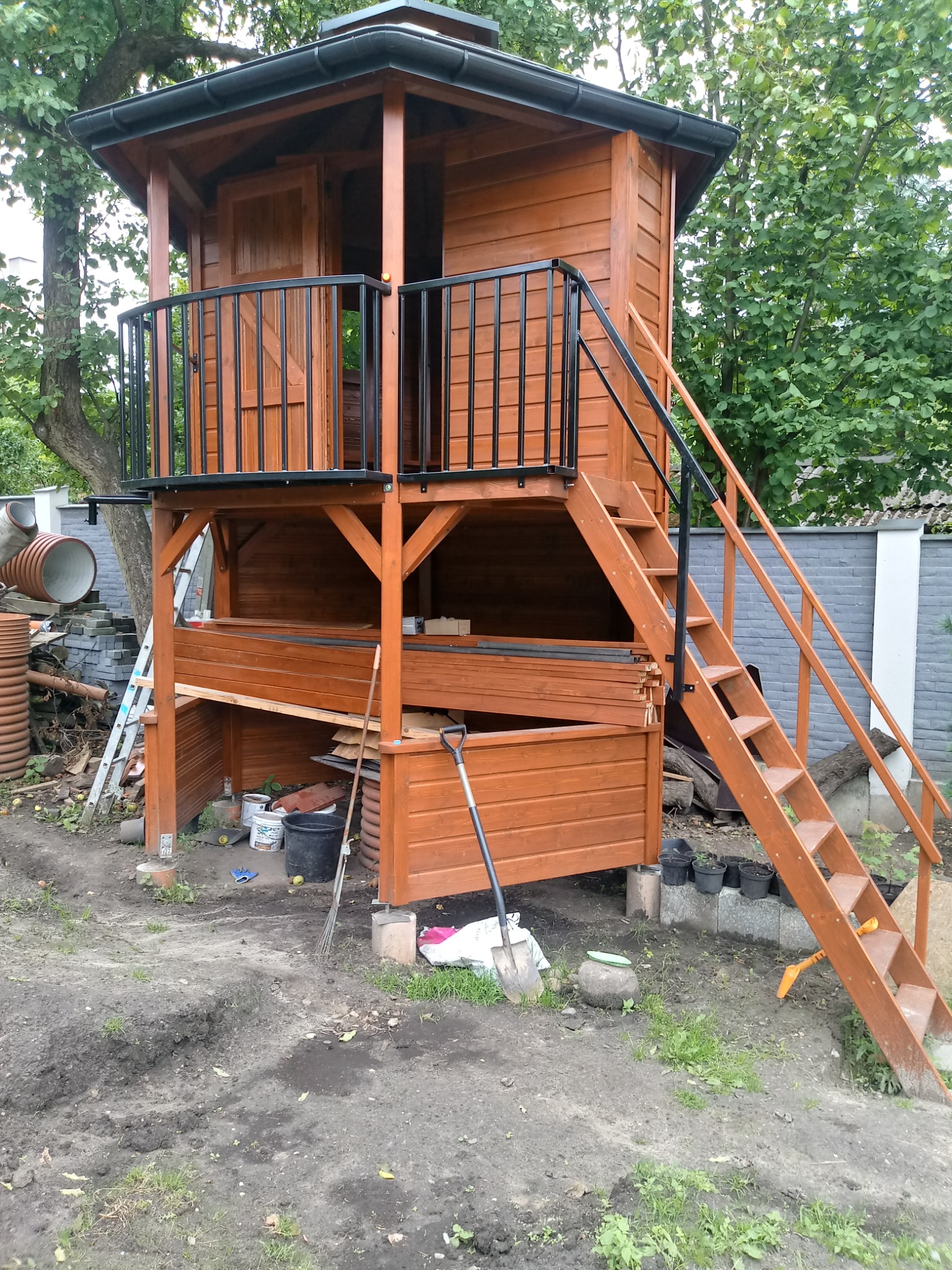 Drewniana konstrukcja domku na palach z balustradą i schodami, widok z zewnątrz, otoczenie w trakcie prac ziemnych, widoczne narzędzia ogrodnicze i materiały budowlane.