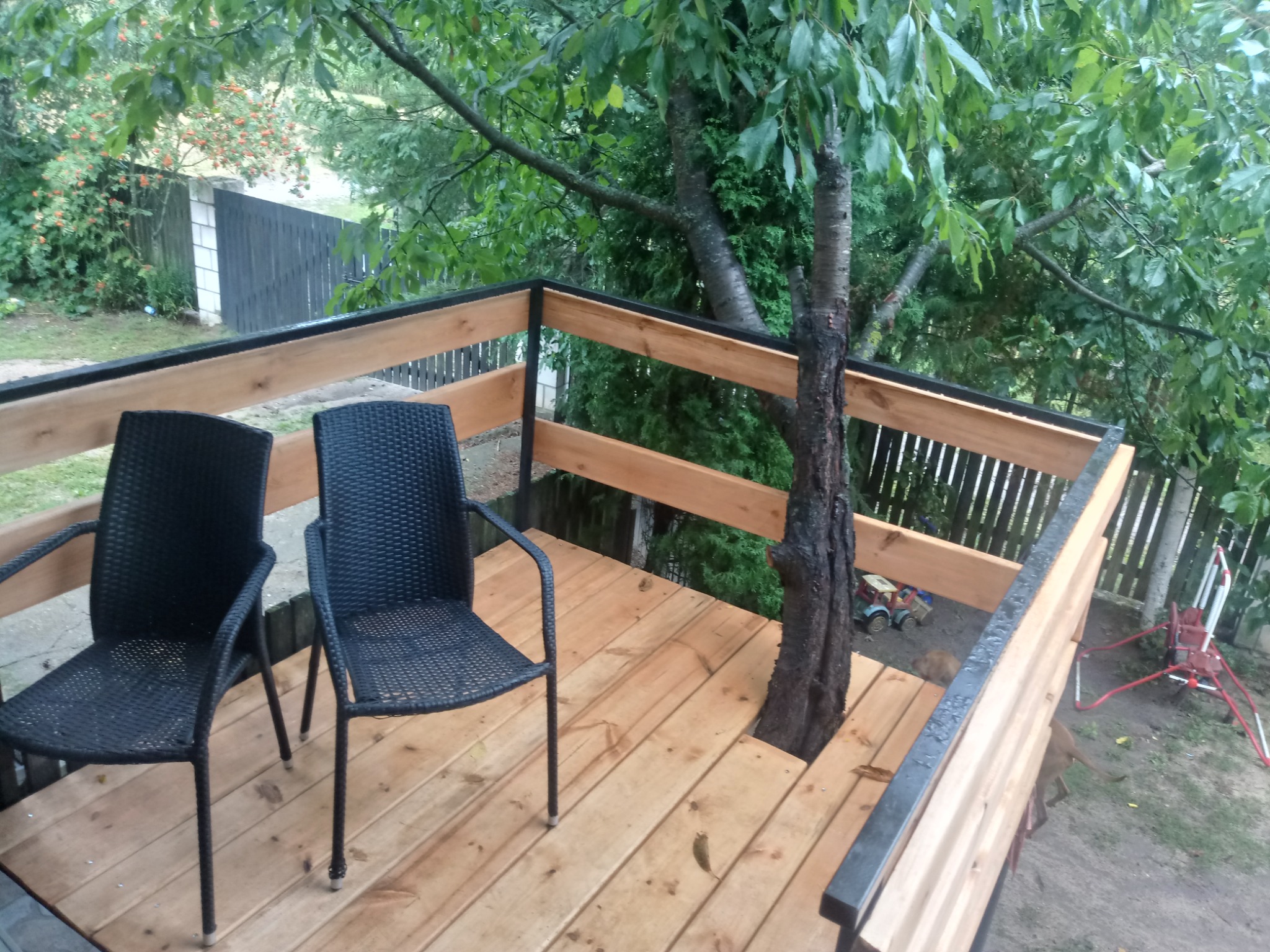 Drewniany taras z balustradą zintegrowany z drzewem, wyposażony w dwa czarne krzesła z plecionki, widok na ogród z zabawkami.