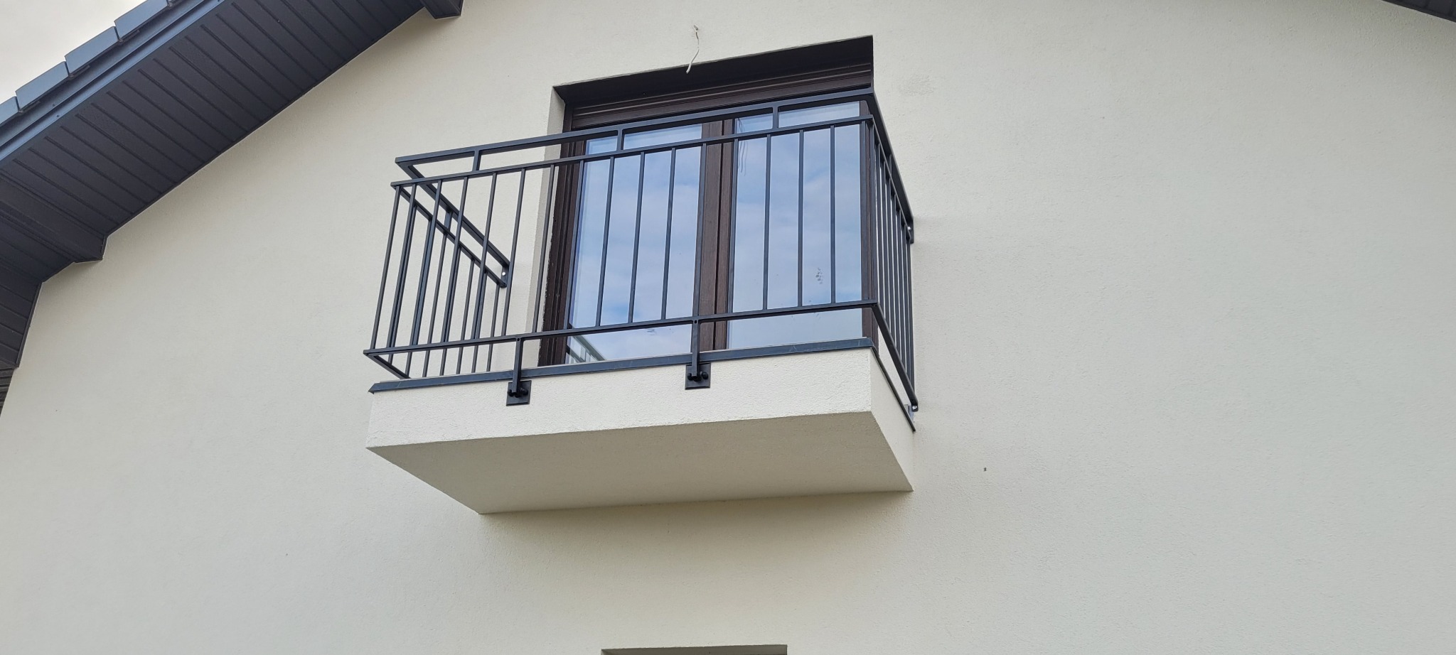 Czarna, metalowa balustrada balkonowa z pionowymi prętami, zamocowana do jasnej elewacji budynku z ciemną stolarką okienną i grafitowym dachem.