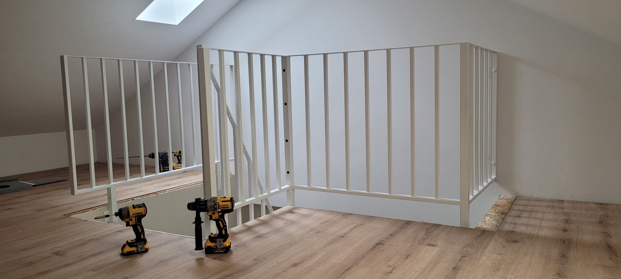 Biała balustrada zabezpieczająca schody na poddaszu, z dwoma wiertarkami akumulatorowymi DeWalt na pierwszym planie i w tle, drewnopodobna podłoga, okno dachowe.