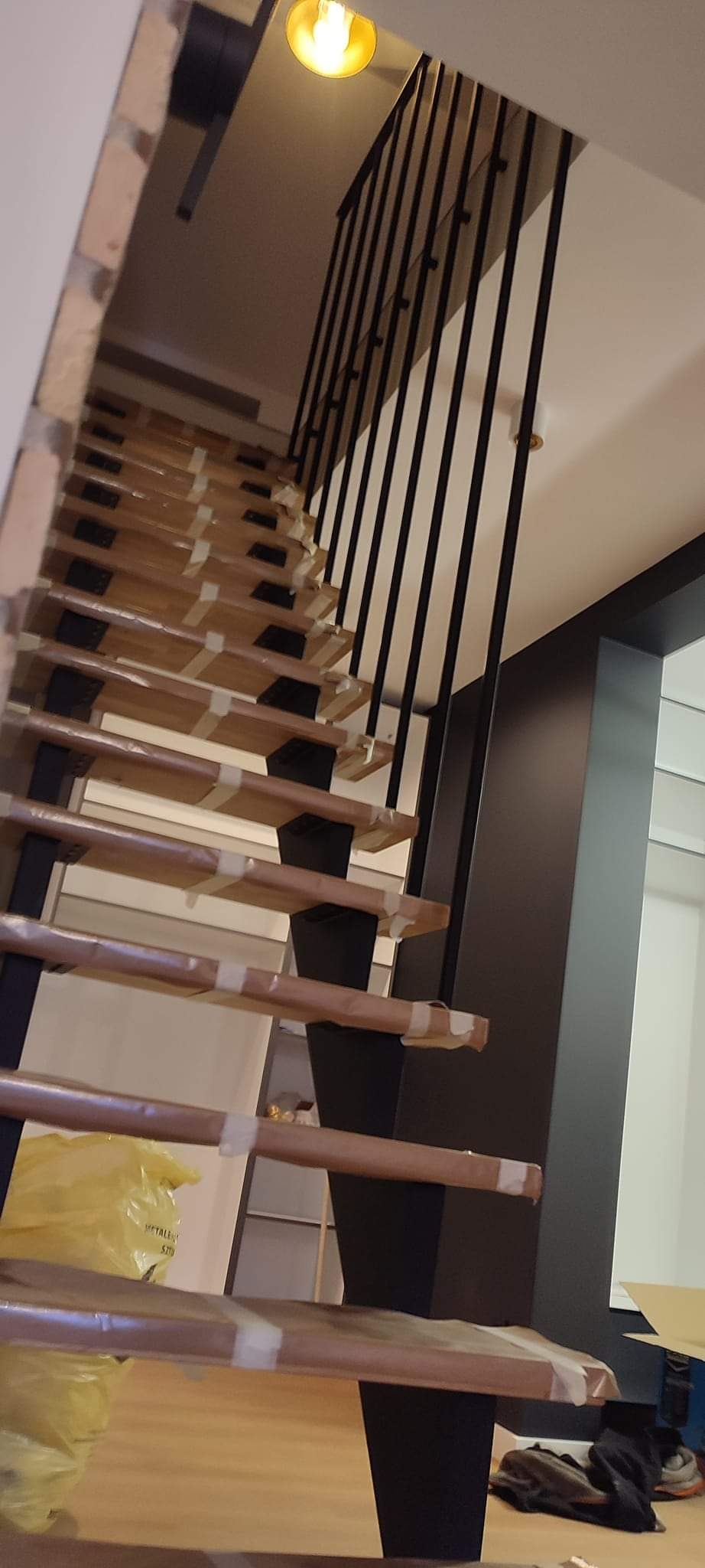Wnętrze z drewnianymi schodami z czarną, metalową balustradą, zabezpieczonymi folią i taśmą na czas montażu, widok od dołu.
